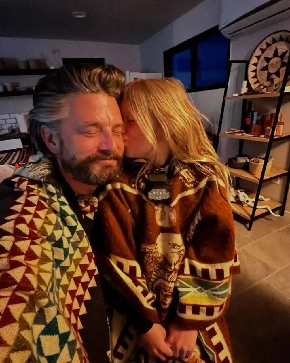 Indiana Elle kisses her father, Jac Cheairs, on the cheek while they both wear vibrant, patterned blankets in a cozy room.